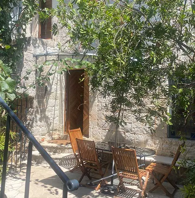 Steinterrasse mit Holzmöbeln im Schatten von Bäumen vor einem traditionellen Natursteinhaus.
