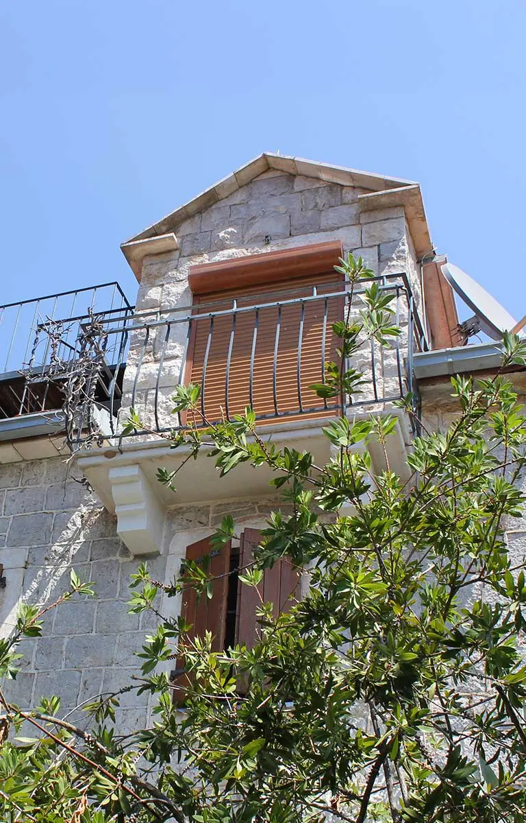 Steinernes Ferienhaus mit Balkon, Holzläden und mediterraner Begrünung unter blauem Himmel.