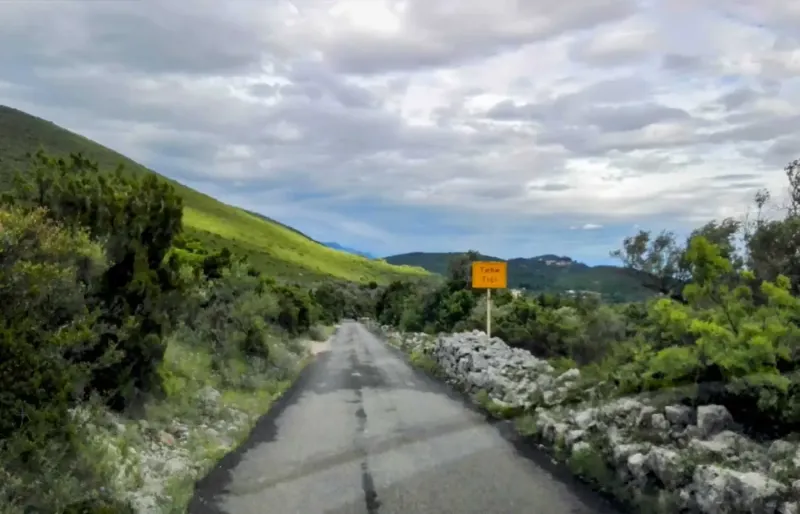 Narrow country road through green hills with village sign in distance